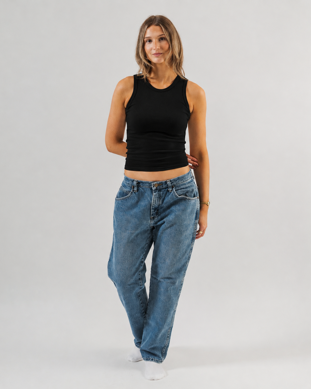 A woman modeling a black sleeveless crop top and loose blue jeans against a light gray studio background. - Women's Basics - Baby Rib Tank Top
