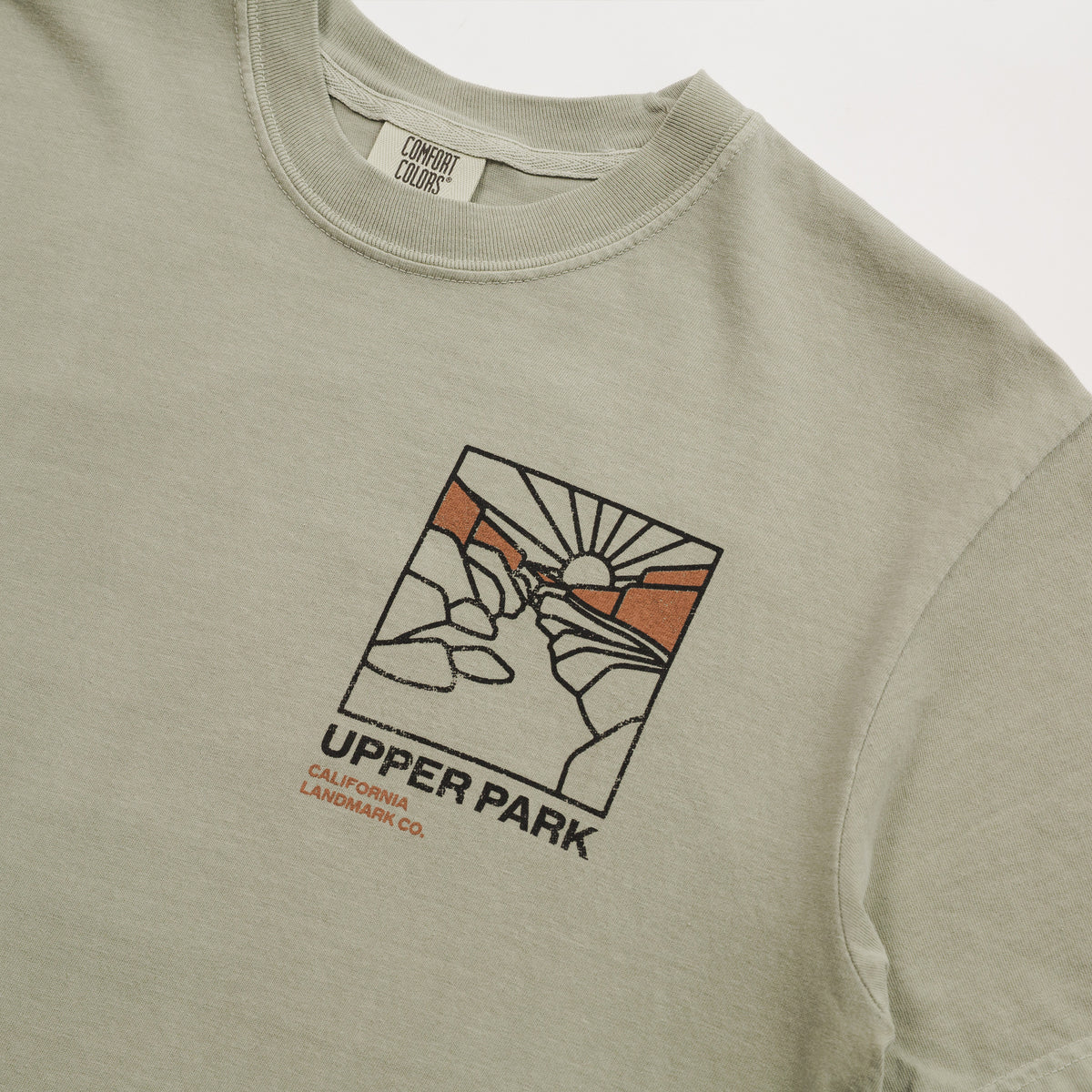 Close-up of a light sage green t-shirt featuring a graphic print of a hand holding a pen with a sun and mountains, with the text 'UPPER PARK CALIFORNIA LANDMARK CO.' printed below.