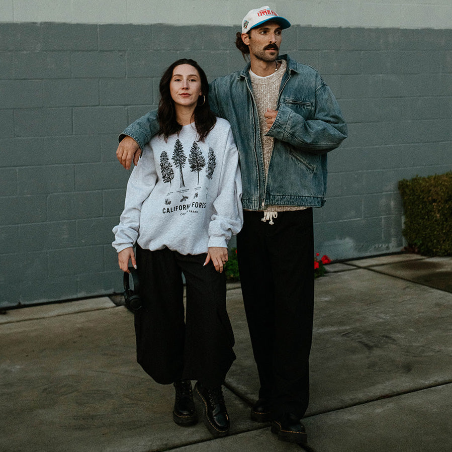 Two people standing against a gray wall, one wearing a white sweatshirt with tree graphics and the other in a denim jacket. Upper Park - California Forests Crew Neck sweatshirt