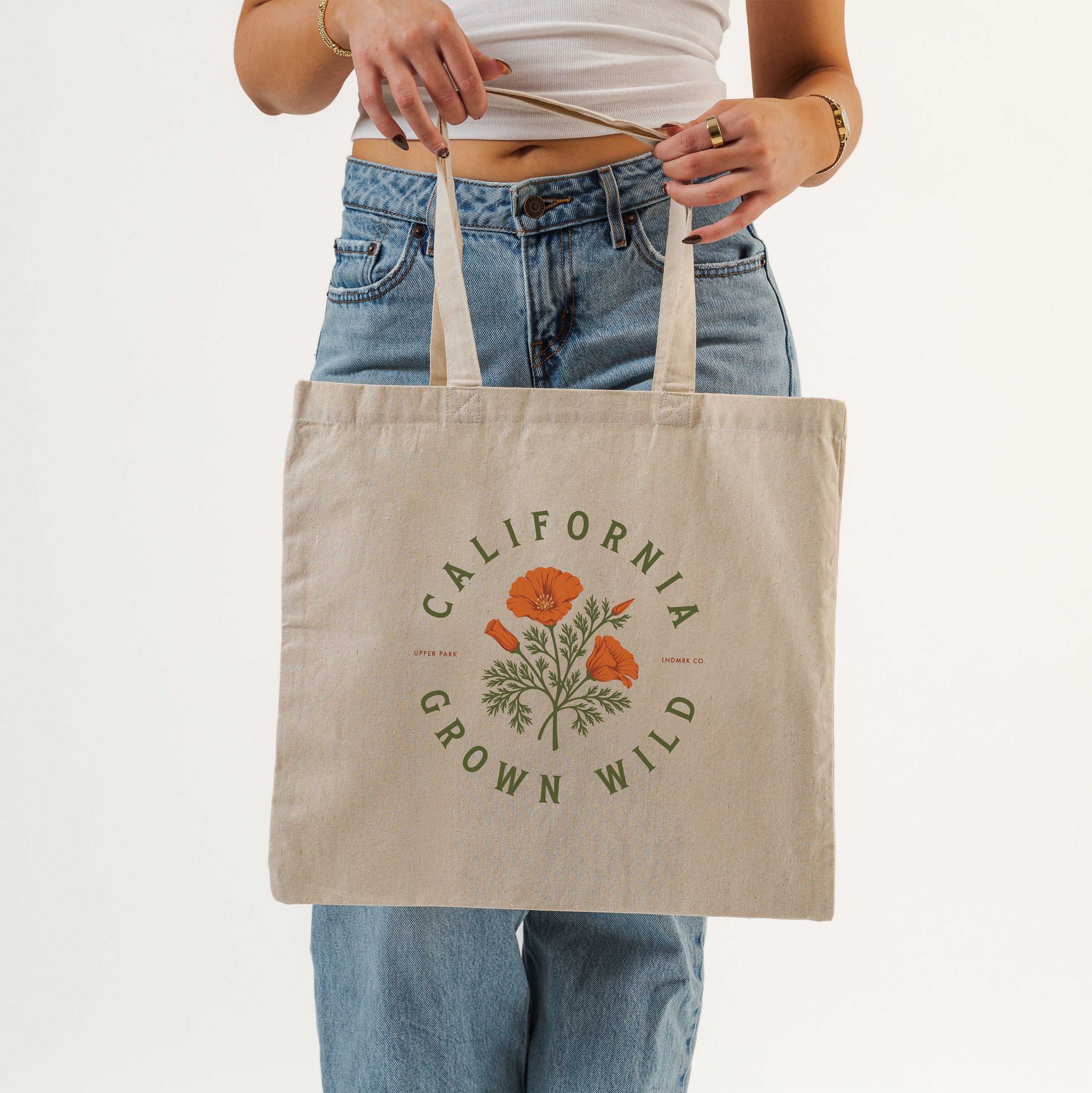 A person wearing a white crop top and blue jeans holds a beige canvas tote bag with a green and orange poppy graphic and the text 'CALIFORNIA GROWN WILD'.