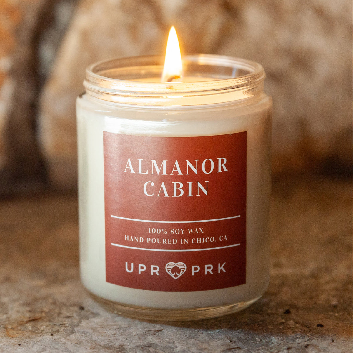 An 8oz soy wax candle jar with a label that reads 'Almanor Cabin', lit with a visible flame, placed on a stone surface.