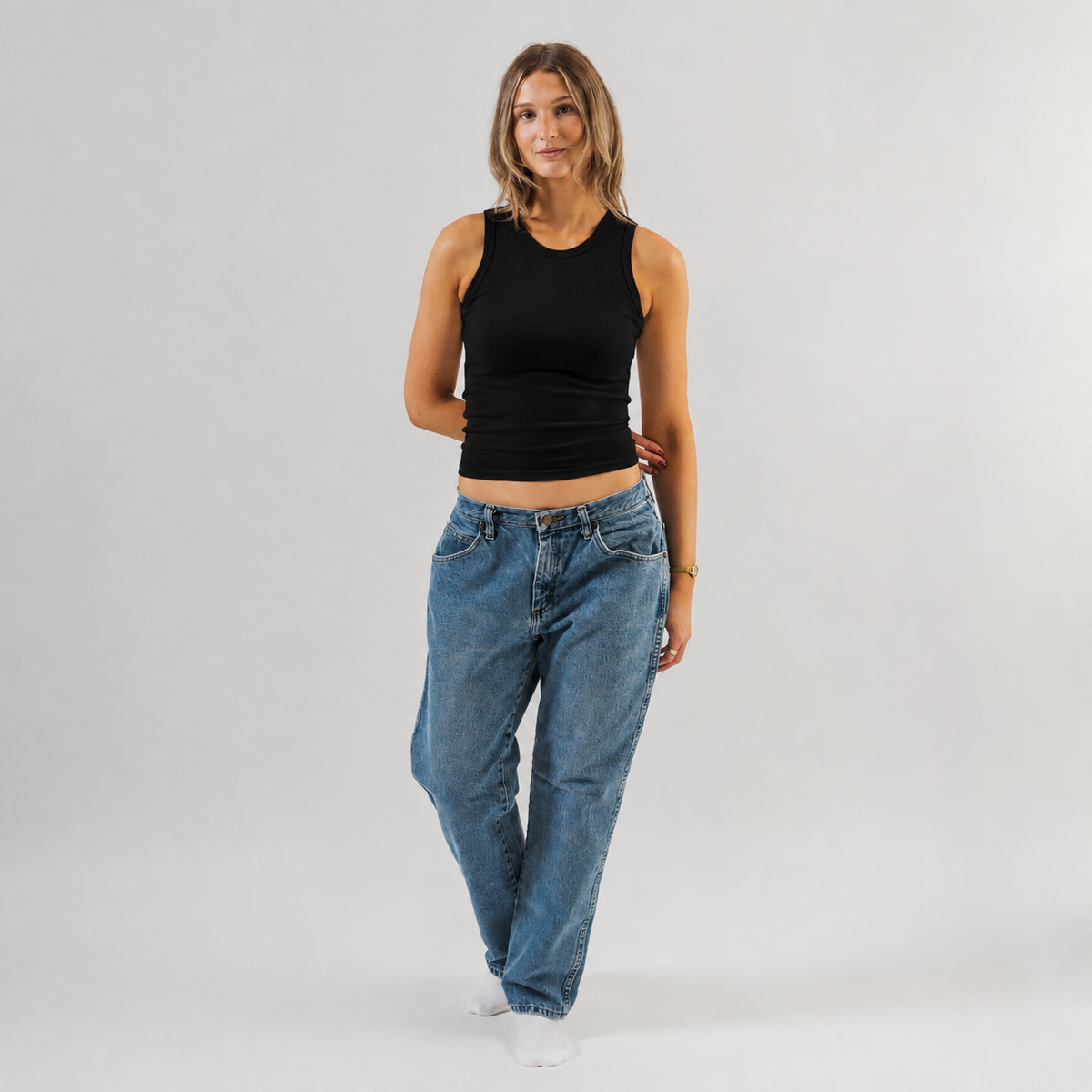 A woman modeling a black sleeveless crop top and loose blue jeans against a light gray studio background.  - Women's Basics - Baby Rib Tank Top