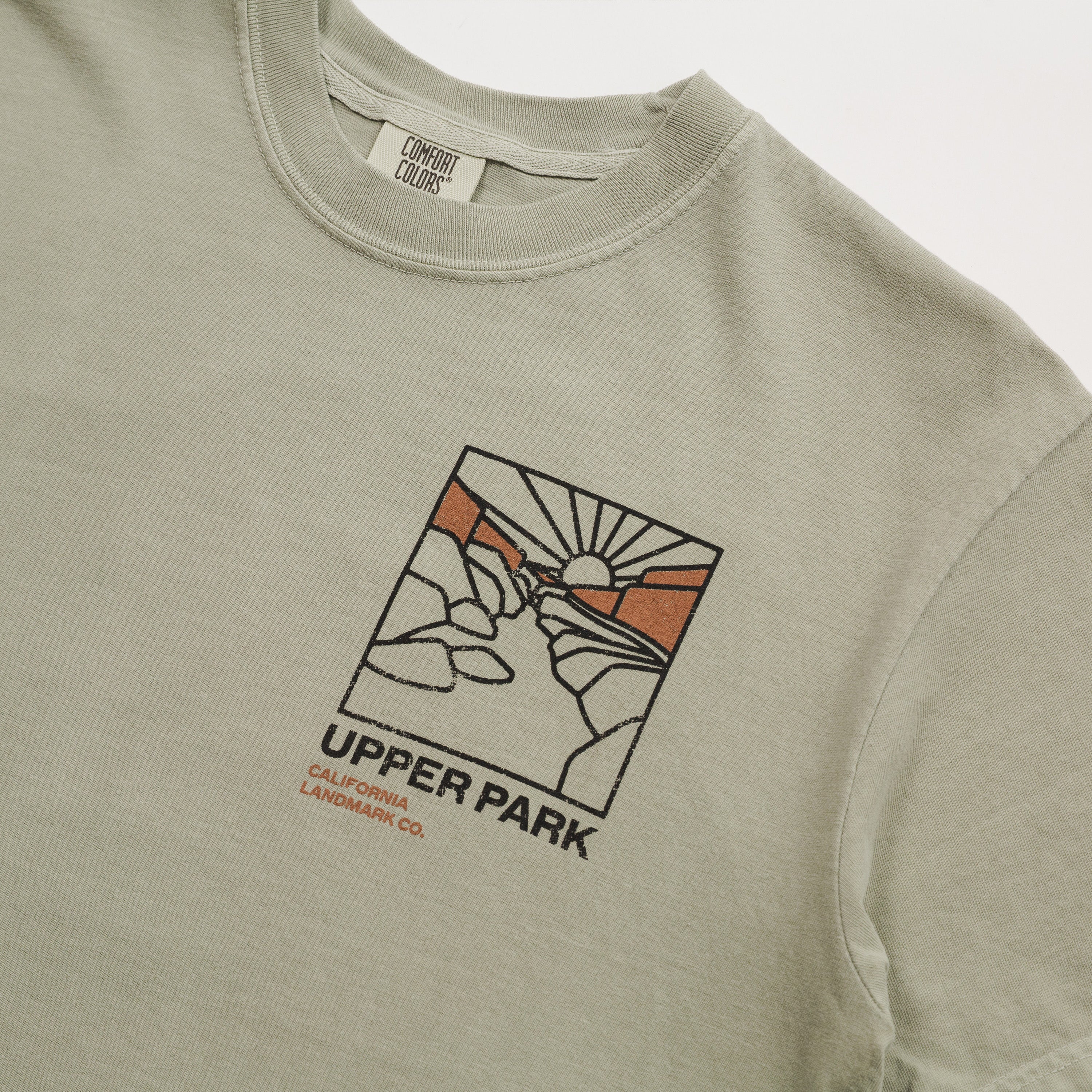 Close-up of a light sage green t-shirt featuring a graphic print of a hand holding a pen with a sun and mountains, with the text 'UPPER PARK CALIFORNIA LANDMARK CO.' printed below.