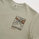 Close-up of a light sage green t-shirt featuring a graphic print of a hand holding a pen with a sun and mountains, with the text 'UPPER PARK CALIFORNIA LANDMARK CO.' printed below.
