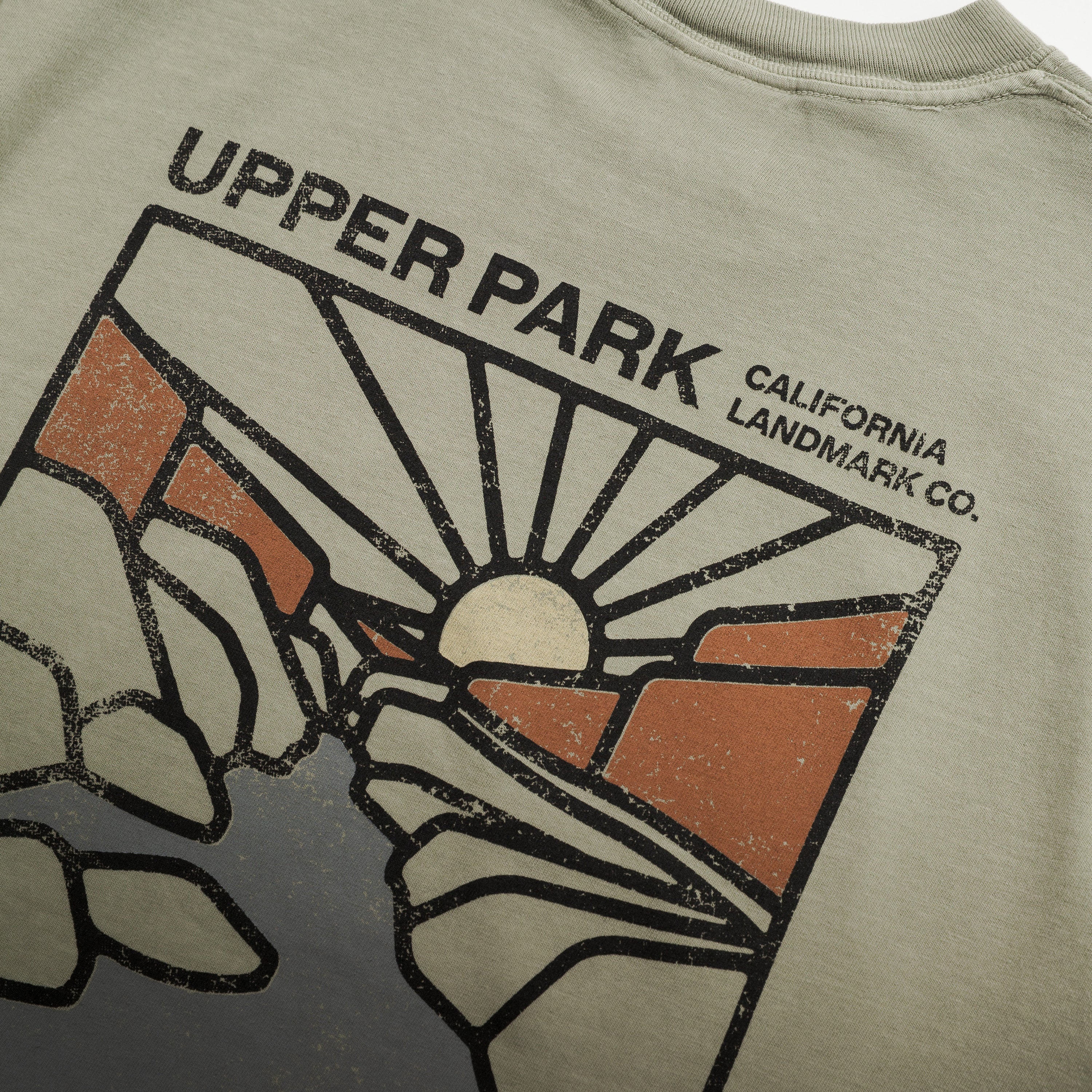 Close-up of a vintage-style graphic t-shirt showing the back print with 'UPPER PARK CALIFORNIA LANDMARK CO.' text and a stylized sun-and-rock design in black, orange, and off-white on a light beige fabric.