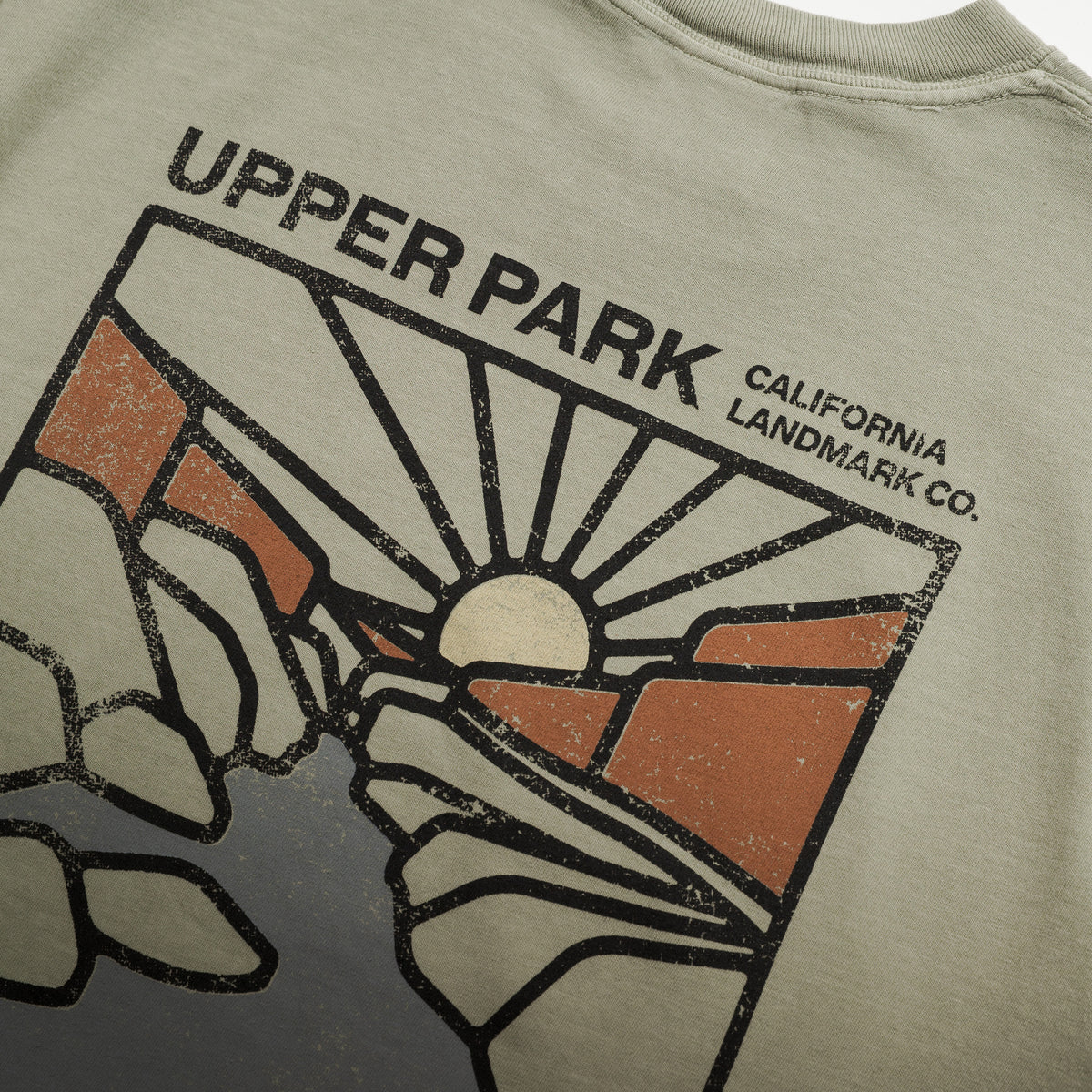 Close-up of a vintage-style graphic t-shirt showing the back print with 'UPPER PARK CALIFORNIA LANDMARK CO.' text and a stylized sun-and-rock design in black, orange, and off-white on a light beige fabric.
