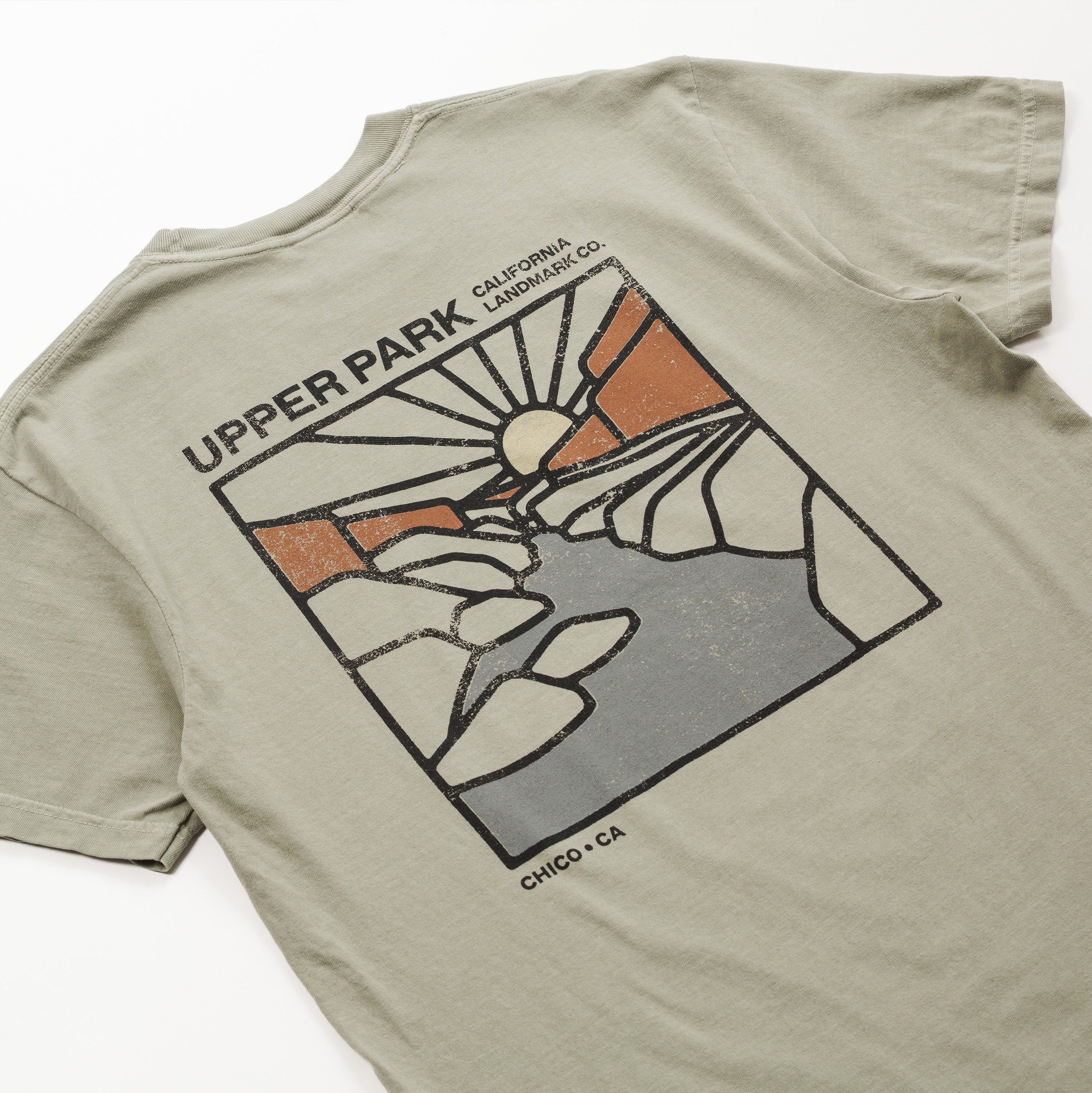 A close-up of the back of a light olive green t-shirt featuring a distressed graphic of Upper Park, California, with a stylized sun, mountains, and rocks in orange, gray, and black.