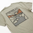 A close-up of the back of a light olive green t-shirt featuring a distressed graphic of Upper Park, California, with a stylized sun, mountains, and rocks in orange, gray, and black.