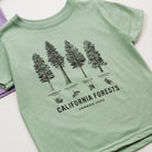 A light sage green short-sleeve t-shirt featuring a black line art graphic of four California forest trees with labels, and the text 'CALIFORNIA FORESTS COMMON TREES' printed below.