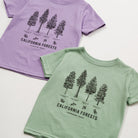 Two short-sleeve kids' t-shirts laid flat on a white background, one in light purple and one in light sage green, both featuring a black line art graphic of California forests with tree illustrations and text.