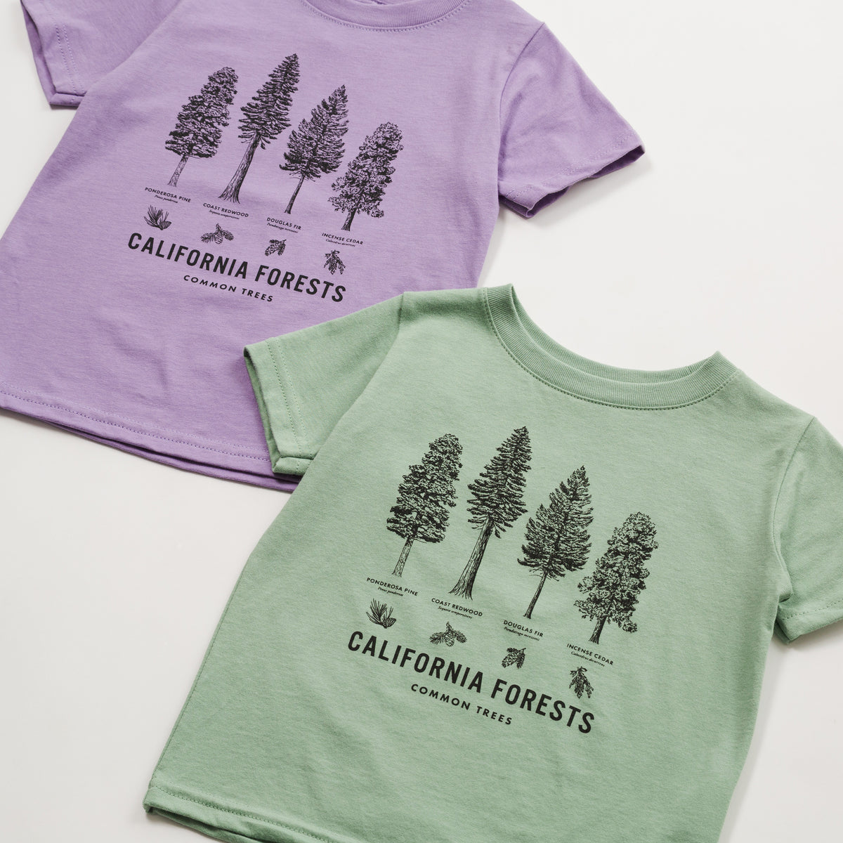 Two short-sleeve kids' t-shirts laid flat on a white background, one in light purple and one in light sage green, both featuring a black line art graphic of California forests with tree illustrations and text.
