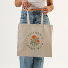 A person wearing a white crop top and blue jeans holds a beige canvas tote bag with a green and orange poppy graphic and the text 'CALIFORNIA GROWN WILD'.