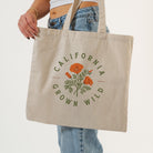 A person wearing blue jeans holds a natural canvas tote bag with a green and orange California Grown Wild graphic print, against a clean white background.