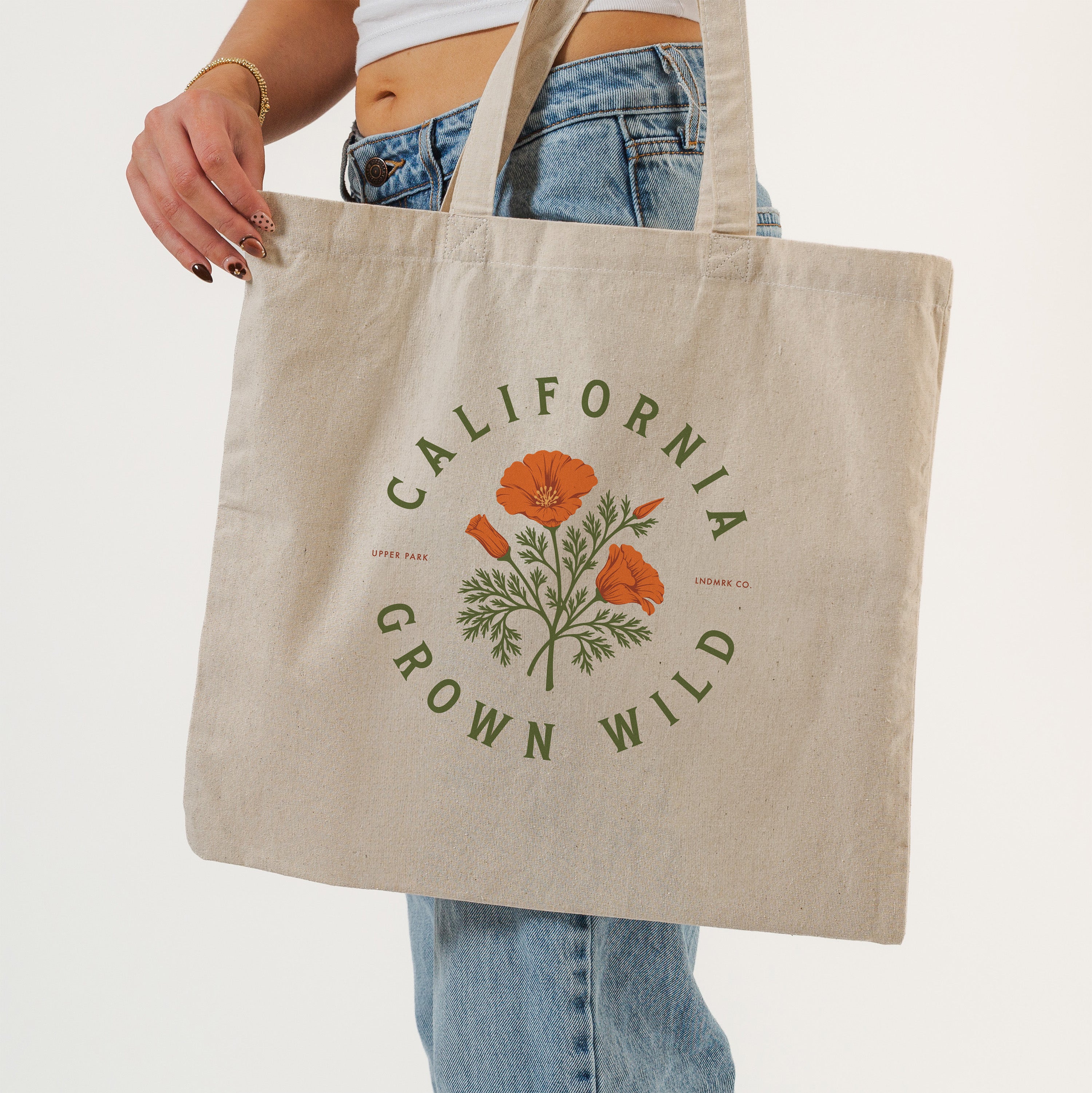 A person wearing blue jeans holds a natural canvas tote bag with a green and orange California Grown Wild graphic print, against a clean white background.