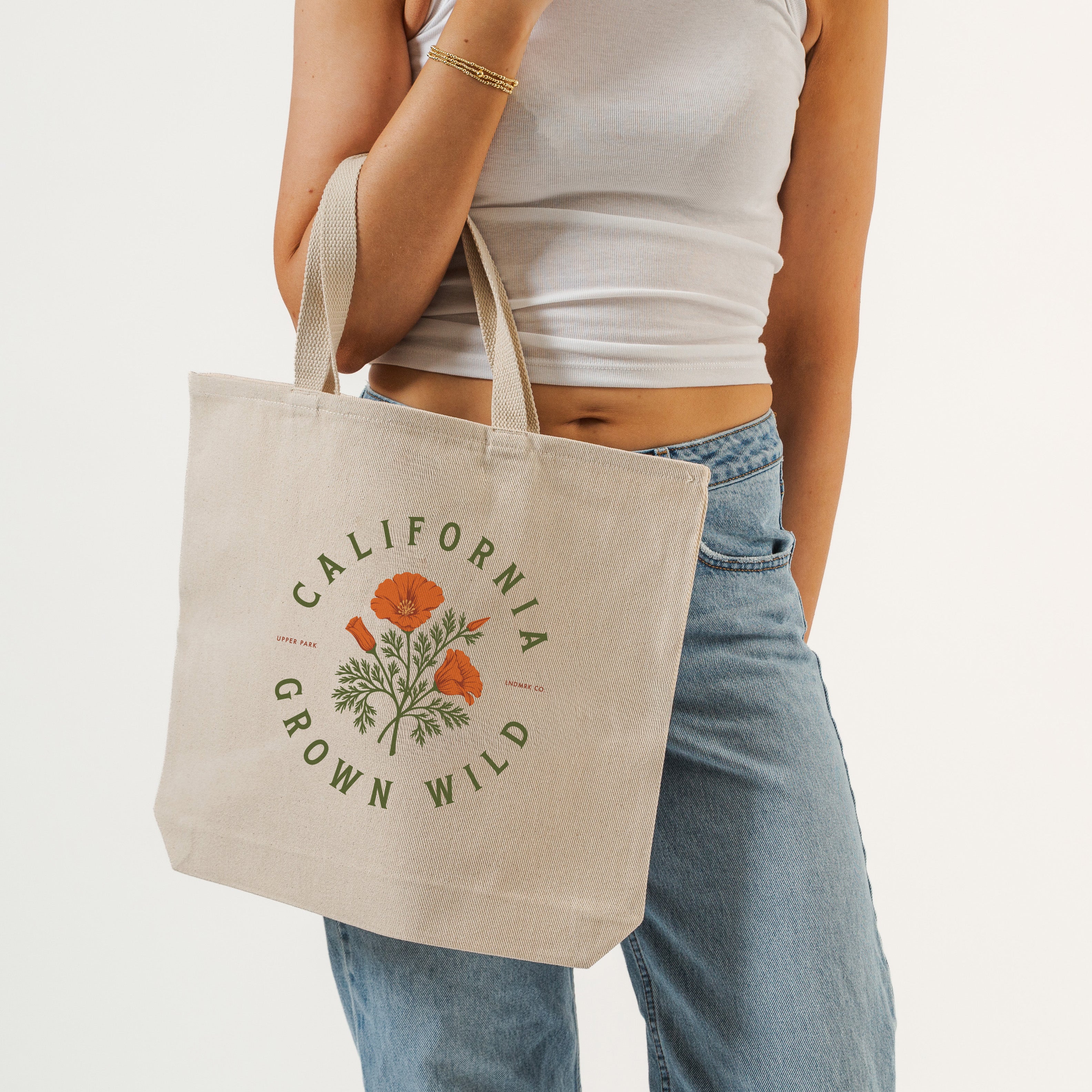 A person wearing a white ribbed tank top and light blue jeans carries a natural canvas tote bag. The bag features an orange poppy floral illustration and the text 'CALIFORNIA GROWN WILD' printed in olive green.
