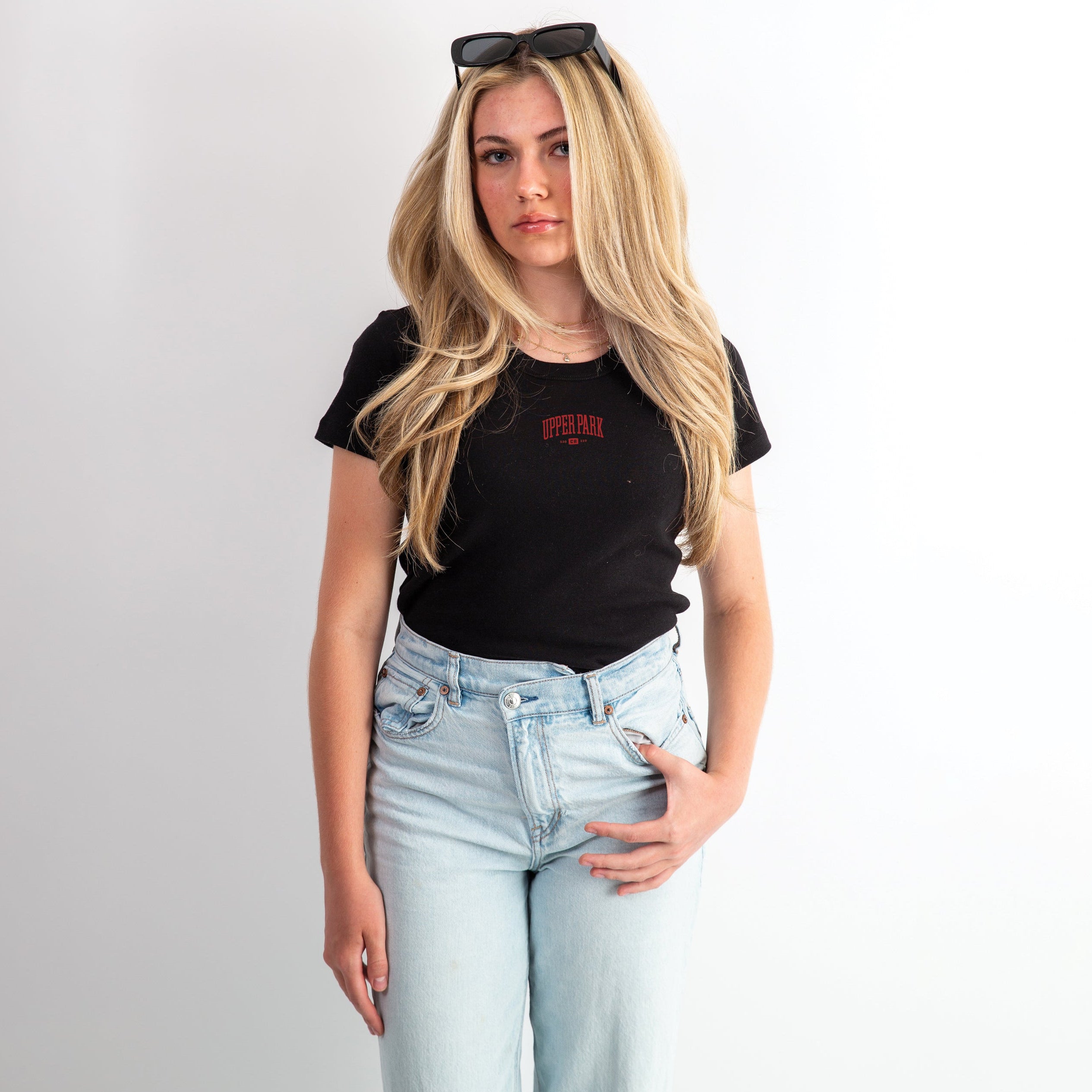 A woman wearing a black organic rib baby tee with a university-inspired design, paired with light blue jeans and sunglasses on her head.