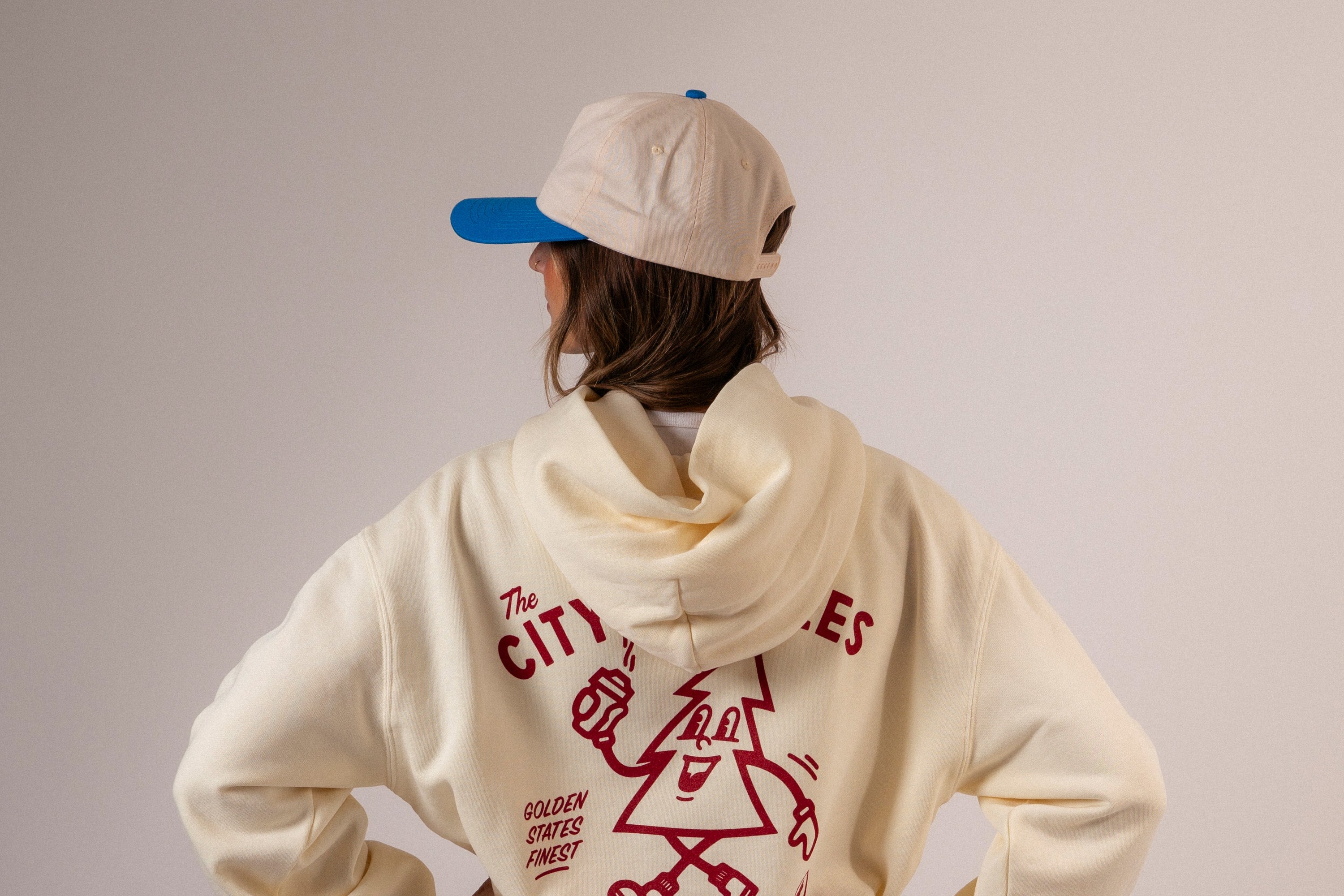 Person wearing a cream hoodie with red text and graphics, standing against a plain background. Upper Park Clothing Hats Banner