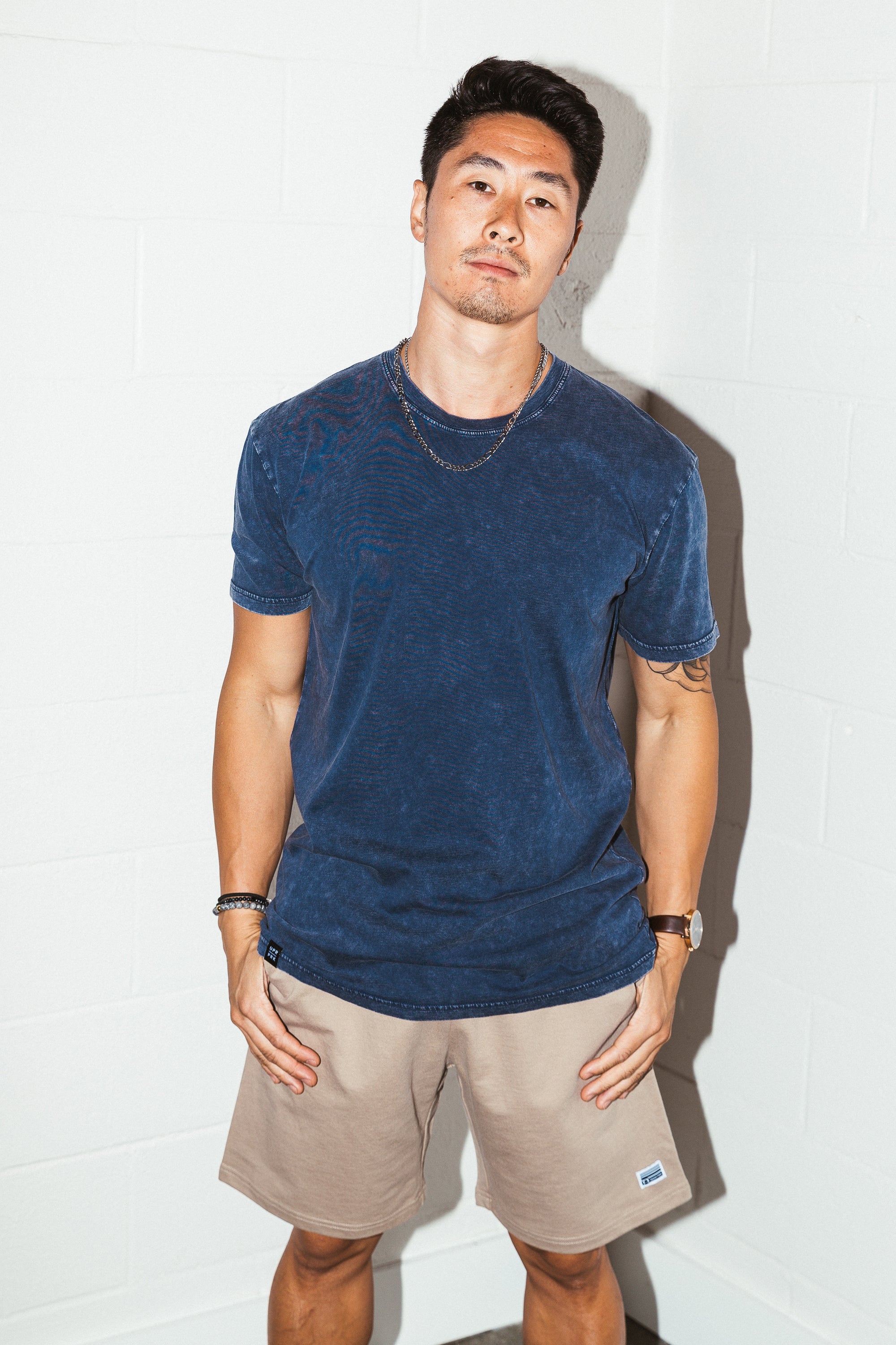 man leaning against a white brick wall taken with flash photography wearing a Simple Stone Washed Blue Tee with khaki colored mens sweat shorts from Upper Park Clothing in Chico, California