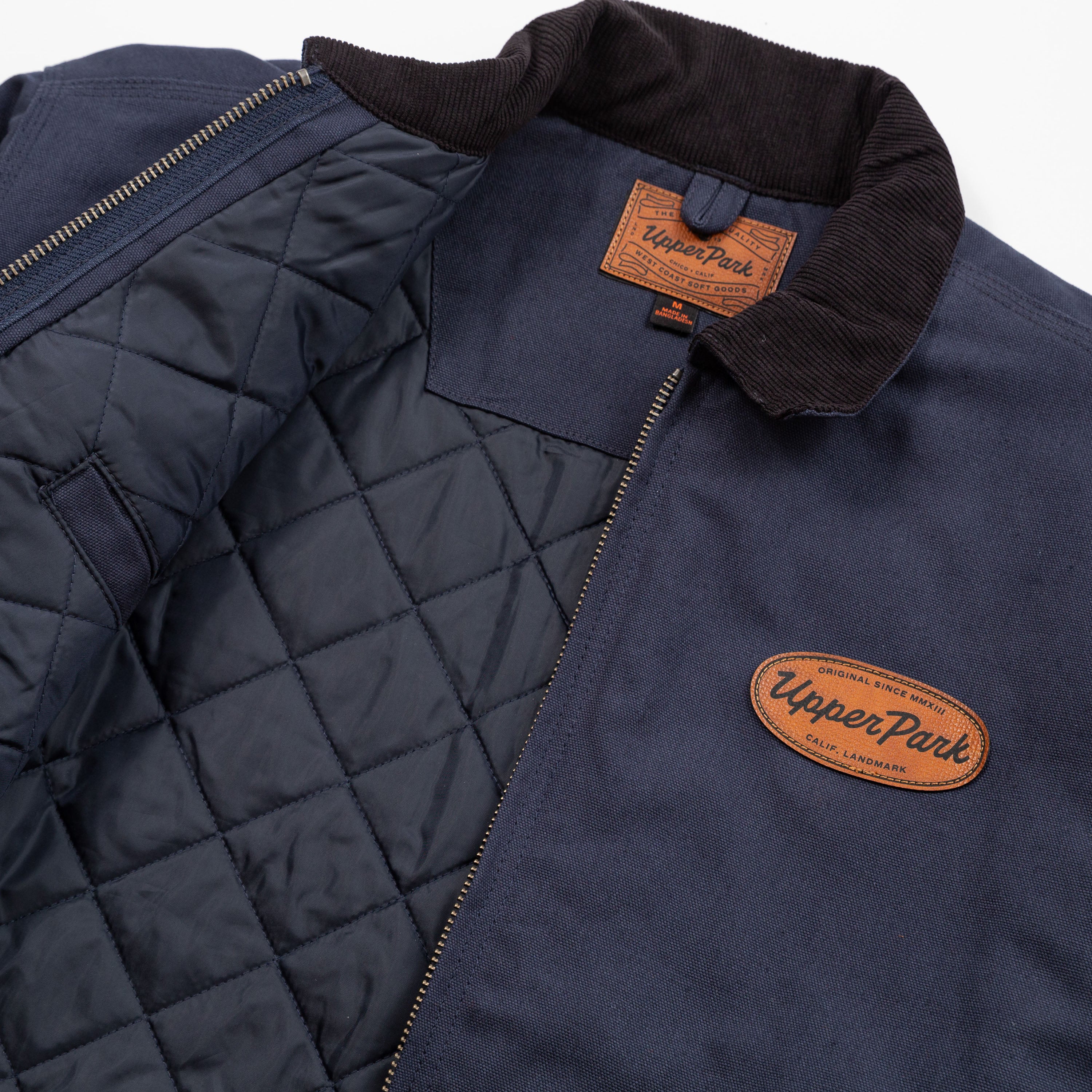 Navy quilted jacket with brown leather patches displaying 'Upper Park' brand on a white background - Cultivators Upper Park Barn Jacket