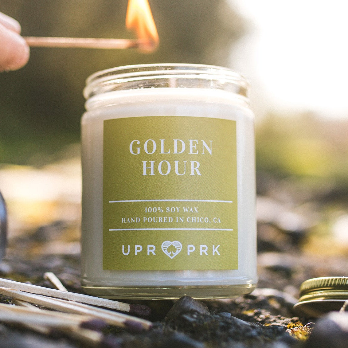 An 8oz jar candle with a label reading 'GOLDEN HOUR' being lit outdoors, part of a sunset picnic setup.